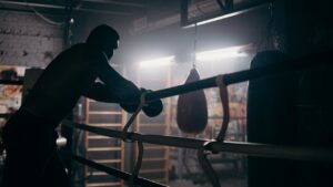 Bokser leunt vermoeid op de touwen in een donkere gym met een boksbal op de achtergrond