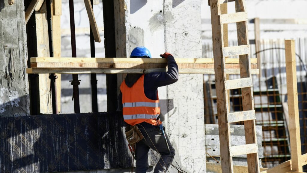 Bouwvakker met helm en oranje hesje draagt houten balk op bouwplaats voor aanbouw, focus op kosten aanbouw.